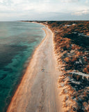 Aerial view of a long sandy beach curving along the left side of turquoise water, with dunes and shrubs on the right under a pale cloudy sky.