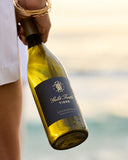 A hand holding a bottle of Gold Turtle Tides Chardonnay, photographed against a softly blurred beach background at sunset.