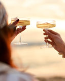 Two hands holding up champagne coupe glasses filled with a golden beverage, clinking together in a toast against a warm sunset background.