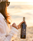 A person in a white jacket and sunglasses holds a bottle of Gold Turtle Tides Cabernet Sauvignon red wine on a beach at sunset.