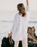 A woman stands on a rocky shoreline with the ocean in the background, wearing an oversized white long-sleeve shirt and light pants while holding a bottle of Gold Turtle Tides Prosecco.