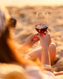 A person sitting on a sandy beach at sunset holds a glass of red wine, with warm golden light and a blurred beach background.