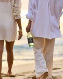 Two people walking on a sandy beach at sunset, viewed from behind. One person holds a bottle of Gold Turtle Tides SSB.